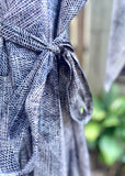 Close-up of a gray and white textured woven fabric tied in a knot, with a blurred outdoor background of green foliage and a wooden fence.