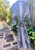 A gray and white patterned long-sleeve wrap robe with a tie waist, hanging on a wooden hanger outdoors on a garden path.