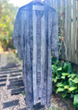 A lightweight kimonohanging outdoors on a hanger, featuring a gray and white abstract pattern. The garment is shown against a blurred background of a wooden fence, green plants, and stone steps.