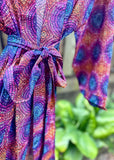 A close-up of a colorful patterned garment with a tie waist, featuring concentric circular dot designs in purple, blue, pink, orange, and red, with a blurred green plant in the background.