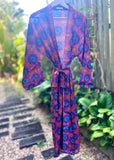 A lightweight wrap-style robe with a colorful mandala pattern in purple, blue, orange, and pink, hanging on a hanger outdoors on a garden path.