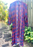 A sheer long-sleeve open-front cover-up dress with a vibrant purple, blue, and orange pattern, hanging on a hanger outdoors on a garden path.