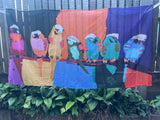 A large rectangular fabric banner or tapestry featuring a colorful painted design of birds perched on a branch, hanging outdoors in front of a fence and green plants. The artwork uses bold blocks of black, orange, purple, blue, yellow, pink, and teal.