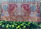 A large square abstract textile or wall hanging with a dense dot pattern in earthy tones, suspended outdoors in front of a fence and green plants. The design features symmetrical organic shapes in burgundy, tan, brown, cream, and light blue.