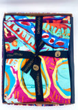 A folded tablecloth presented in an open box, tied with a black and gold ribbon.